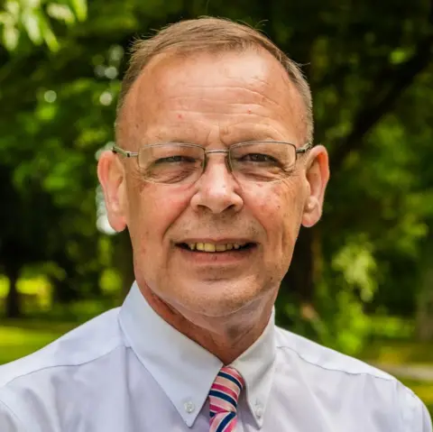 Anthony Lloyd A man with short brown hair has rectangular thin-rimmed glasses on and is smiling for a head and shoulders shot. He is wearing a white shirt with a pink and blue stripy tie and trees can be seen blurred behind him.