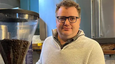 Julia Gregory/BBC Oliver Leach - a man with glasses and a beige fleece looking into the camera. He is standing in a kitchen with a coffee machine with beans in next to him.