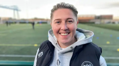 Abbey Lyle smiling at the camera with the football pitch behind her. She is wearing a grey sweatshirt and a black body warmer.  She has short blonde which is tied back.