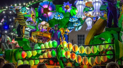Simon Boddy A stunning carnival float of neon green, yellow and blue flowers, garlands, leaves and men in purple suits. The Walls logo is visible on the float