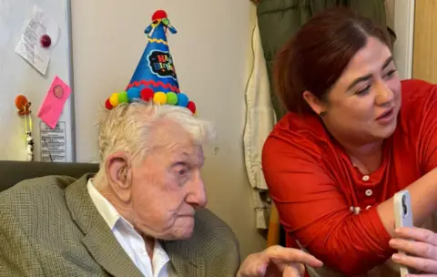 An elderly man in a colourful party hat looks at a mobile phone held by a care home worker  