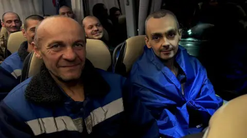Zelenskiy/Official via Telgram Two newly-released male prisoners of war, in blue jackets, smile at a camera while sat on a coach