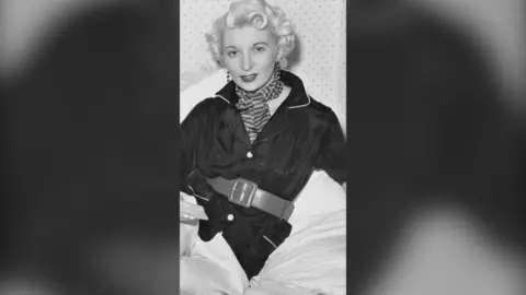 A black and white photo of Ruth Ellis. She has curly blonde hair and is wearing a dark coloured jacket and a striped scarf.