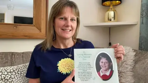 Diane Taylor A woman holding of a card with a photo of a young boy in school uniform printed on it