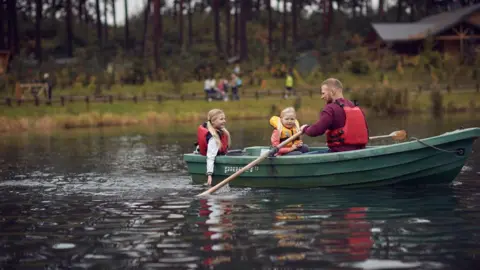 Center Parcs Two children successful  a vessel  with a man, each  wearing lifejackets, connected  a pond with a grassy slope  successful  the background