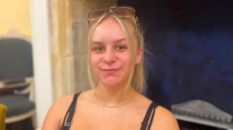 Kelci Hill-Meldrum sits in a chair. She has long blonde hair tied up and a pair of sunglasses propped on the top of her head.