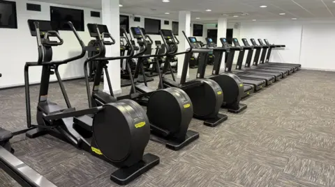 A row of brand new black fitness machines in a gym. To the forefront are four elliptical machines, behind are eight treadmills. There are also various other machines, with large TV screens on the walls.