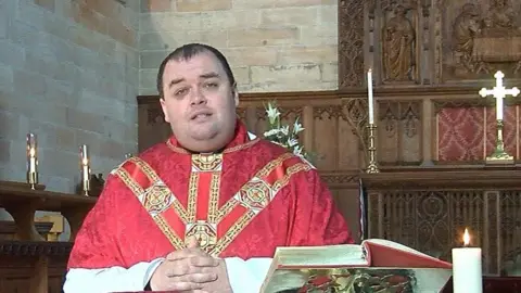 BBC Samuel Erlandson in white, read and gold robes. He at a church pulpit with his hands folded in front of him. there's a thick hardback book on the pedestal in front of him, a burning white candle and a silver cross behind him 