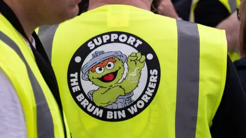 Getty Images A close-up shows the back of a fluorescent yellow hi-vis vest with a large circular badge reading 'Support the Brum Bin Workers' alongside a Unite the Union logo. The badge features a cartoon character emerging from a dustbin with its lid askew. Other people wearing similar hi-vis vests are partially visible on either side.