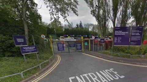 The entrance to Shaw Lane Household Waste Recycling Centre, which is secured with gates. There is purple signage with white writing indicating the site's hours of operation and rules for its use.