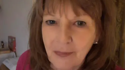 A woman with shoulder-length, light brown hair taking a selfie. She is wearing a red top and is stood in a white room