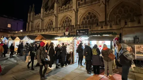 Shoppers are gathered around different stalls at Bath Christmas Market. It is the evening and people are queueing. 