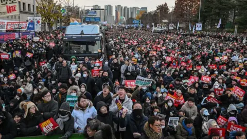 Getty Images People take part in a protest calling for the ouster of South Korean President Yoon Suk Yeol outside the National Assembly in Seoul on December 7, 2024. Huge crowds in winter clothing stand with posters in hand and waving the Korean flag. In the middle is a dark van, likely a police vehicle.