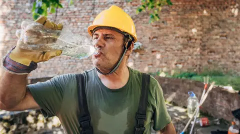 Getty Images Man wearing a yellow safety helmet and sweaty green T-shirt drinking water from a plastic bottle