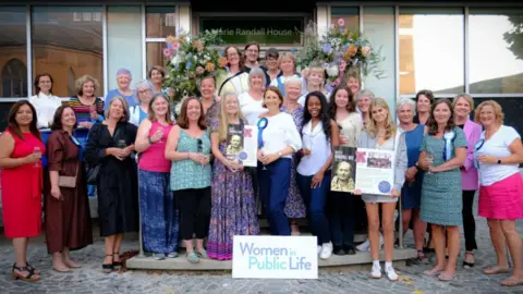 A group of women standing outside Marie Randall House. The group are standing behind a Women in Public Life sign.