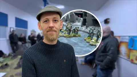 People gather in a room filled with tabletop gaming terrain and miniatures, chatting and watching games unfold. Joe Setterington stands in the foreground while others engage in a lively gaming session in the background.