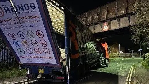 West Mercia Police The back of a lorry that is wedged under a bridge. The back doors and panel of the vehicle is hanging off, and the trailer is caved in.