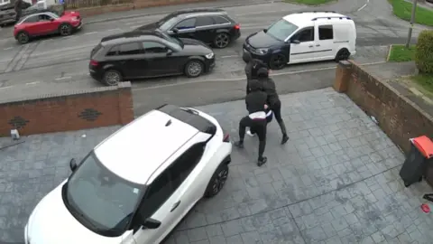 A group of hooded men bundle a man across a driveway towards a black car waiting on the road outside a house.