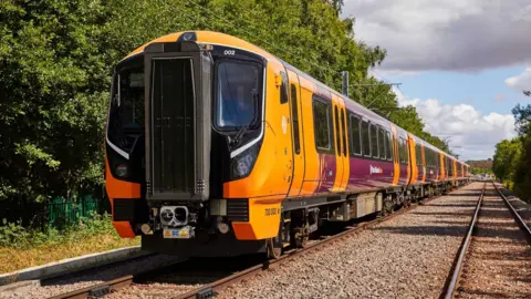 West Midlands Railway A WMR Class 730 train can be seen on tracks. It has several carriages in orange and maroon and trees are visible to the left and right of the tracks. 