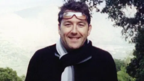 West Yorkshire Police Adam Pickles is standing outside, with trees and mountains behind him. He is wearing a dark coloured top and a white scarf and he is smiling, with glasses worn on his head.