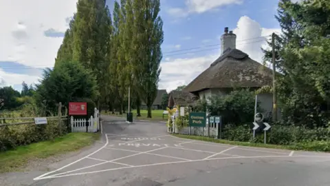 A Google maps image shows the entrance to The Royal Alexandra and Albert School. There are two tall trees in the background. There is a road entrance with a white fence either side. A thatched-roof small cottage sits to the right of the entrance. There are green fields and more trees on the left. 