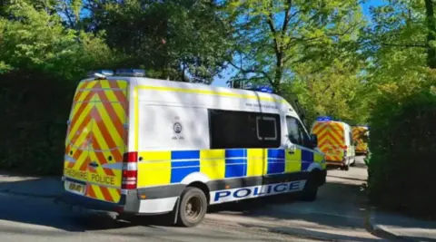 A row of white police vans with blue, yellow and red markings entering a gap in a high hedge