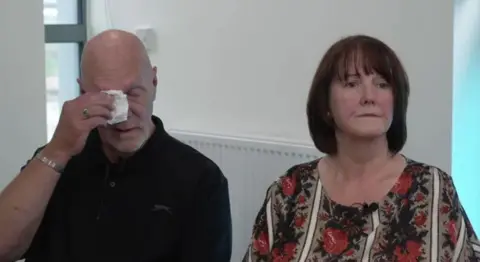 Neil Scott, who is bald and wearing a black polo shirt, wipes his eyes with a tissue as he sits on a chair next to his wife Paula Scott. She has medium-length brown hair and wears a floral blouse, and is looking towards the camera with a solemn expression.