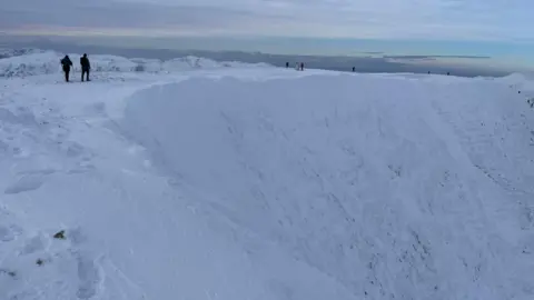 LAKE DISTRICT WEATHERLINE Helvellyn summit in full winter conditions. Snow covers the full summit as two walkers walk into the distance.