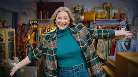 Joanna Page is standing in a charity shop with different kinds of dresses hanging on a rail behind her. There are a number of different items surrounding them in the background. Joanna Page stood infront of them with her arms out wide, smiling at the camera.