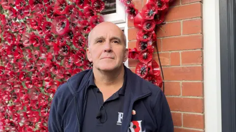A bald gentleman staring at the camera, with a poppy display behind him. He's wearing a navy Royal British Legion Jacket and is wearing a pinned poppy. 