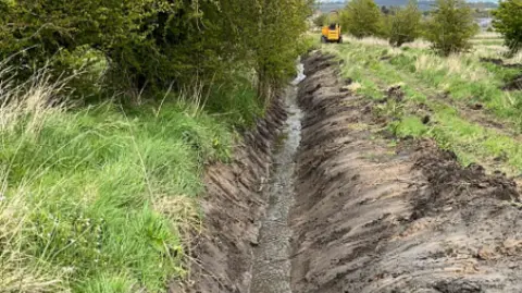 David Wilson A trench with lots of muddy water next to a disused railway track 