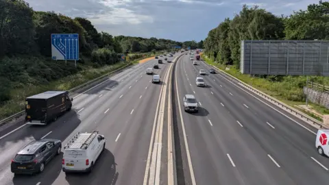 A an aerial shot of a motorway, with traffic on both carriageways. The surface of the road looks new and in good condition. 