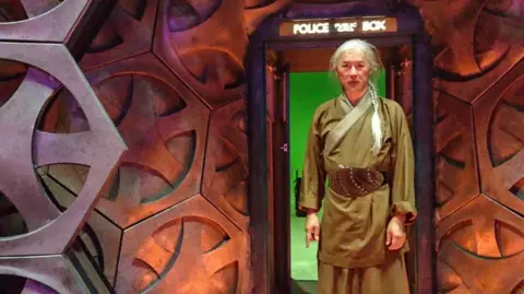 Clem So So has his white long hair in a plait and is wearing a khaki costume. The set looks like a spaceship with a Doctor Who Tardis in the background.