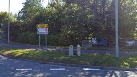 Two stones on a grass area near a roundabout in Liskeard. One of the stones is marked with a 'B'. Signed for Liskeard Community Hospital is next to the stones. Trees are in the background.