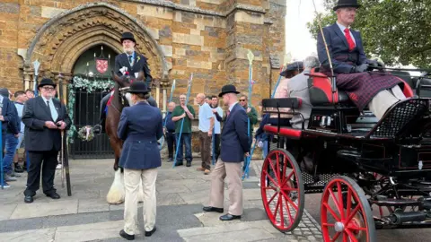 Carroll Weston/BBC A black carriage with red wheels is on the right. On the left is the bailiff on a large brown horse with white feet. He is surrounded by halberdiers – people carrying blue poles.