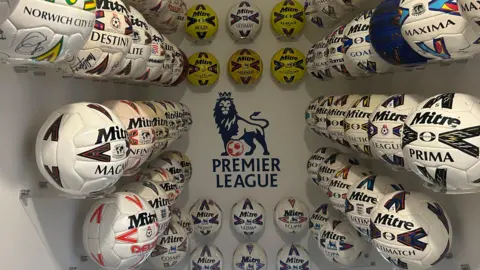A large amount of Mitre footballs mounted and displayed above a staircase on a plain white wall. Each ball has a different Mitre model and design. Stuck at the centre of the back wall is a large Premier League logo.