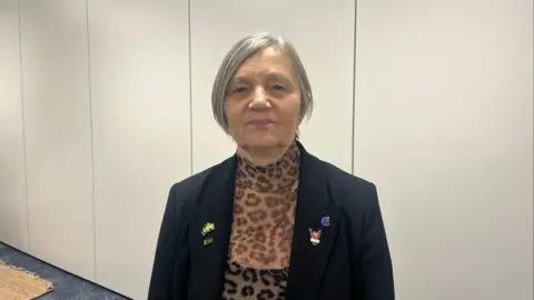 A woman wearing a leopard mess tip and black vest and a black blazer. She has her grey hair tired back and is looking directly at the camera. Behind her is a blank white wall. The carpet is blue. 