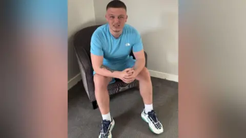 Dale Martin is pictured sitting on a chair wearing a light blue t-shirt, shorts and white trainers.