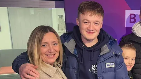 A woman and young man are stood in a radio studio. The man has his arm areound the woman and is dressed in a navy hoodie, blue puffer jacket and has short fair hair. The woman has a short blonde bob and a cream coat. There is a small boy stood behind them, a purple branded display board and a glass window leading to a break out area.