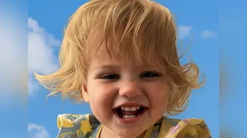 A headshot of a blonde one-year-old girl with a yellow and blue top who is smiling at the camera.
