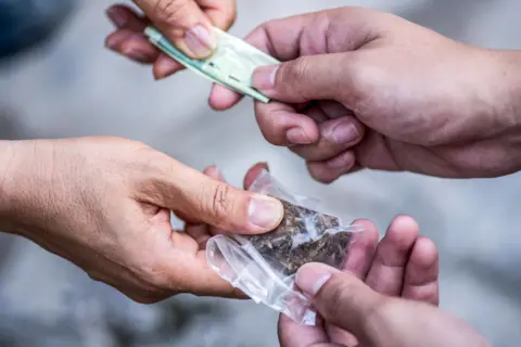 A close up of two sets of hands. A bag of what appears to be cannabis is being exchanged between two of the hands while money is changing hand in the opposite direction