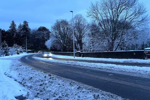 A car driving through a snow-covered street with its lights on.