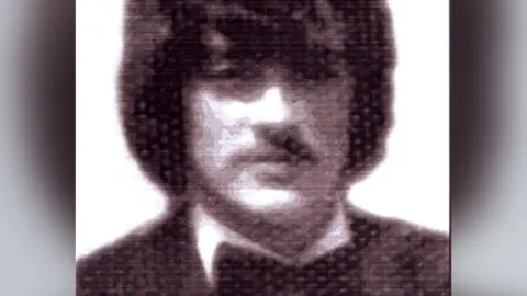 An archive black and white image of Gerard Evans. He is pictured wearing shoulder length dark hair looking directly to camera. He is wearing a dark jacket, white shirt and a black bowtie.