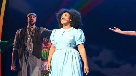 Getty Images Georgina Onuorah on stage at the curtain call of The Wizard of Oz at the London Palladium. She is dressed in a Dorothy costume which is a blue and white gingham dress. She is looking out into the audience ready to take her bow as the rest of the cast looks on, including Ashley Banjo, who is standing behind her to the left.