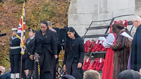 Two women with dark hair and dark coats standing with their heads bowed with women in military uniform to their right, holding a flag and an array of poppy-wreaths to their left