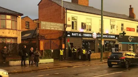 Image shows a street in Balsall Heath, Birmingham, with a few men stood outside a Yemeni restaurant. They are wearing yellow tabards and black hats, and appear to be door staff. 