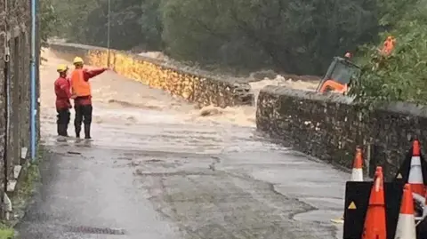 IOM Police Water gushing out of the River Laxey onto the road through a hole in a stone wall. Men in red and orange hi-vis coats and vests point and an orange digger on its side in the river.