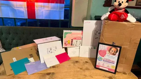Family photograph This picture has a table with lots of cards on it. There is a photo of Ariyah which says "Help us reach 100 birthday cards". A box is on the table with Mini mouse toy sat on top. 