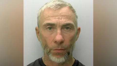 A police mugshot of James Holder. He has cropped grey hair and a short grey beard and is looking directly ahead.