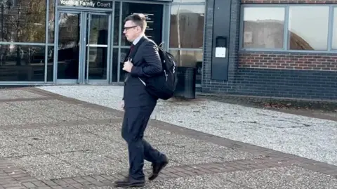 PA Media Christopher Bell walking outside South Tyneside Magistrates' Court on Friday. He is wearing a suit and tie and black-rimmed glasses. he is carrying a backpack. 